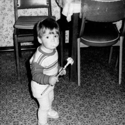 Profilbild Martin. Als Kind zu sehen. Er schaut mit Kuchenteig verschmierten Mund in die Kamera und hält einen Quirl in der Hand. Die am Rande zu sehenden Möbel und Designs entsprechen den 70er Jahren. Das Bild ist mit Schwarz-Weiß Film aufgenommen.